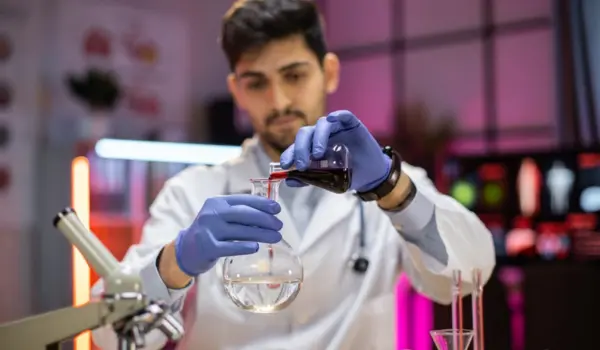 A man in a lab coat carefully pours a colorful liquid from a flask into a clear beaker in a laboratory setting.