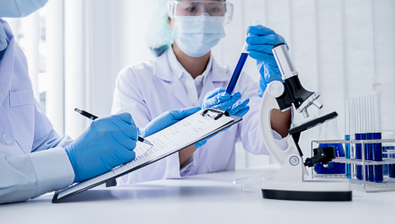 Two individuals in lab coats and masks examine a clipboard, engaged in a discussion in a laboratory setting.